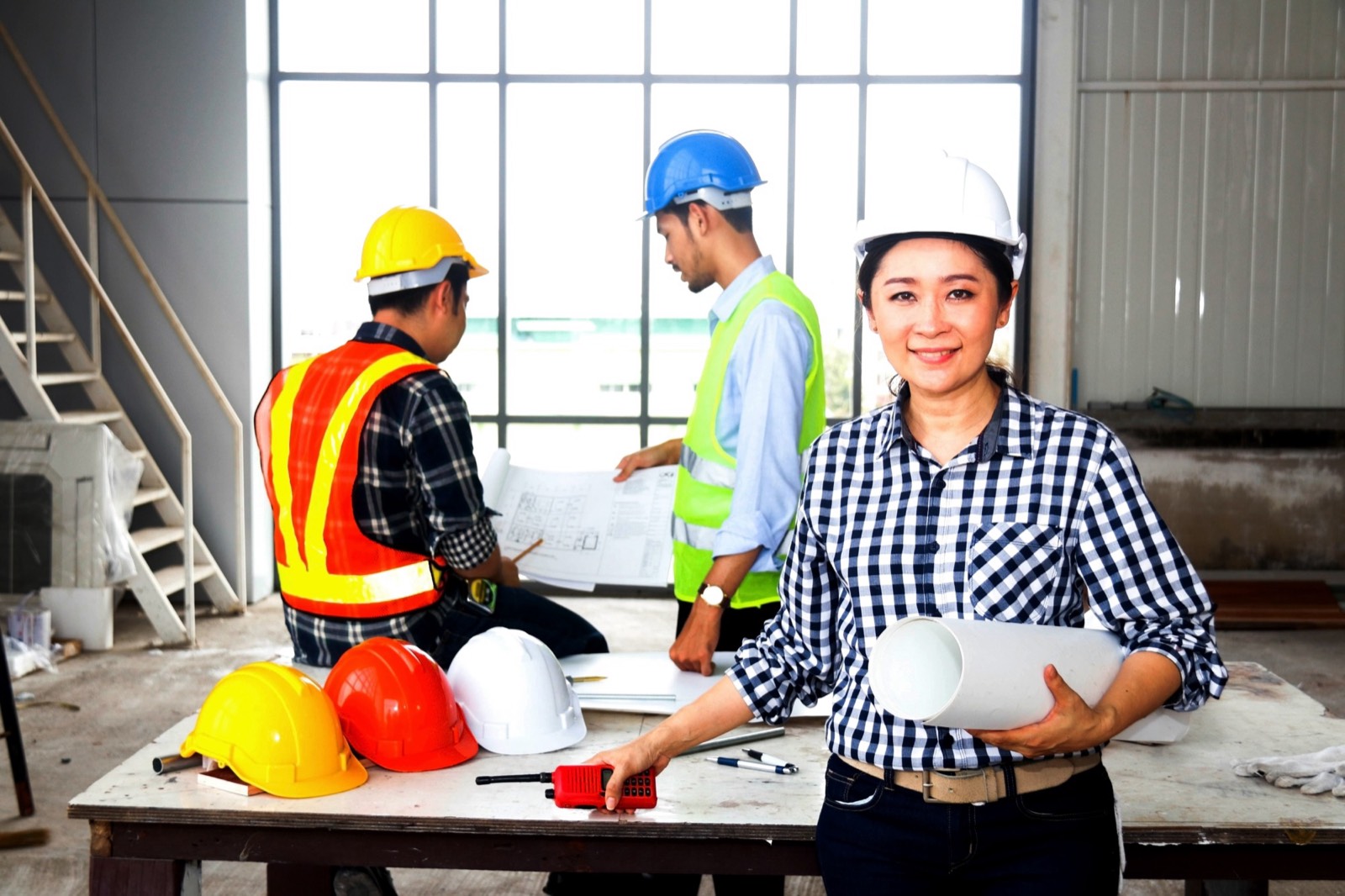 Three people in hard hats around a table with blueprints, one holding a radio